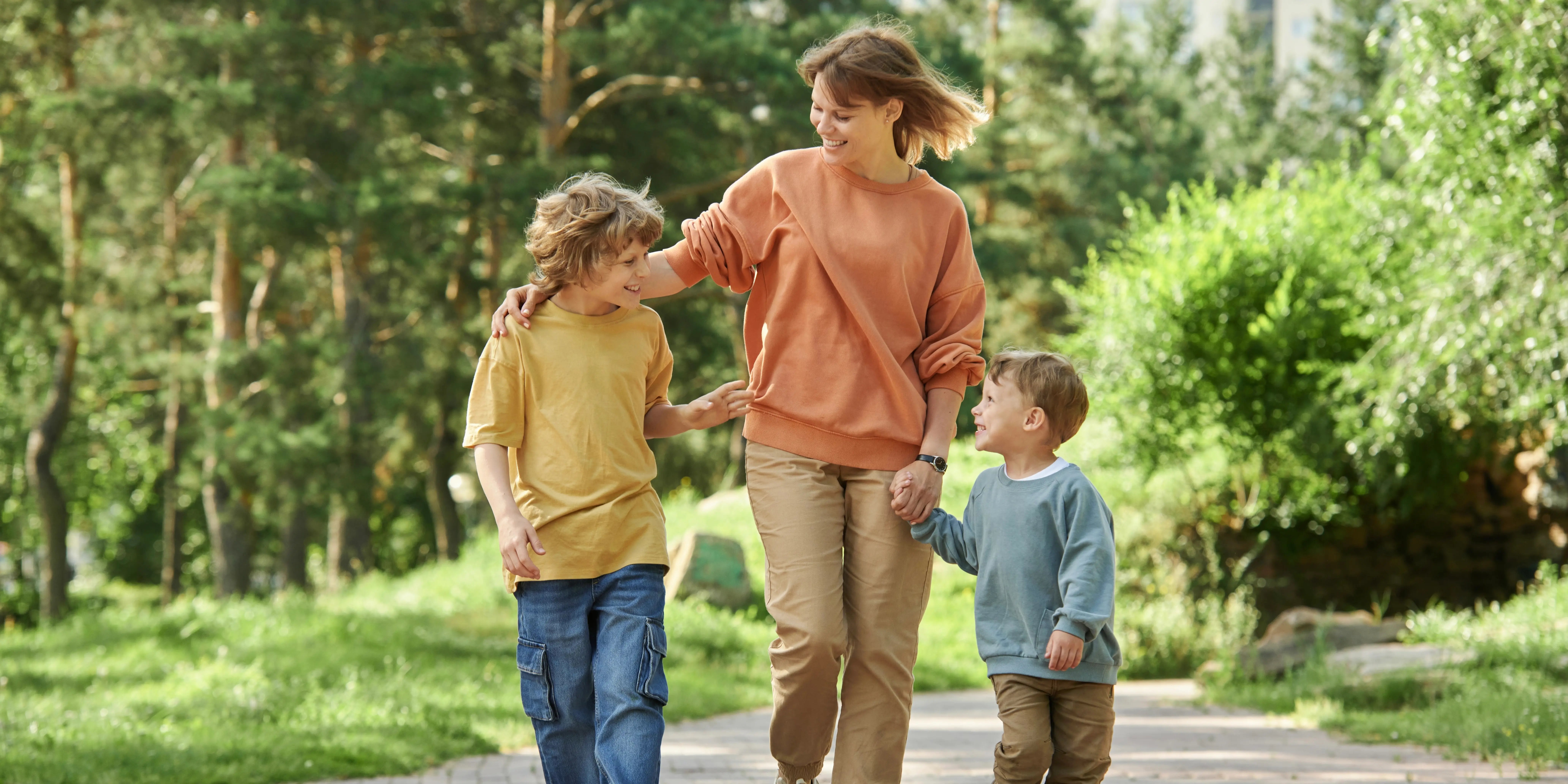 Madre alegre con dos niños tomados de la mano mientras camina en un parque rodeado de árboles en Bosque Real