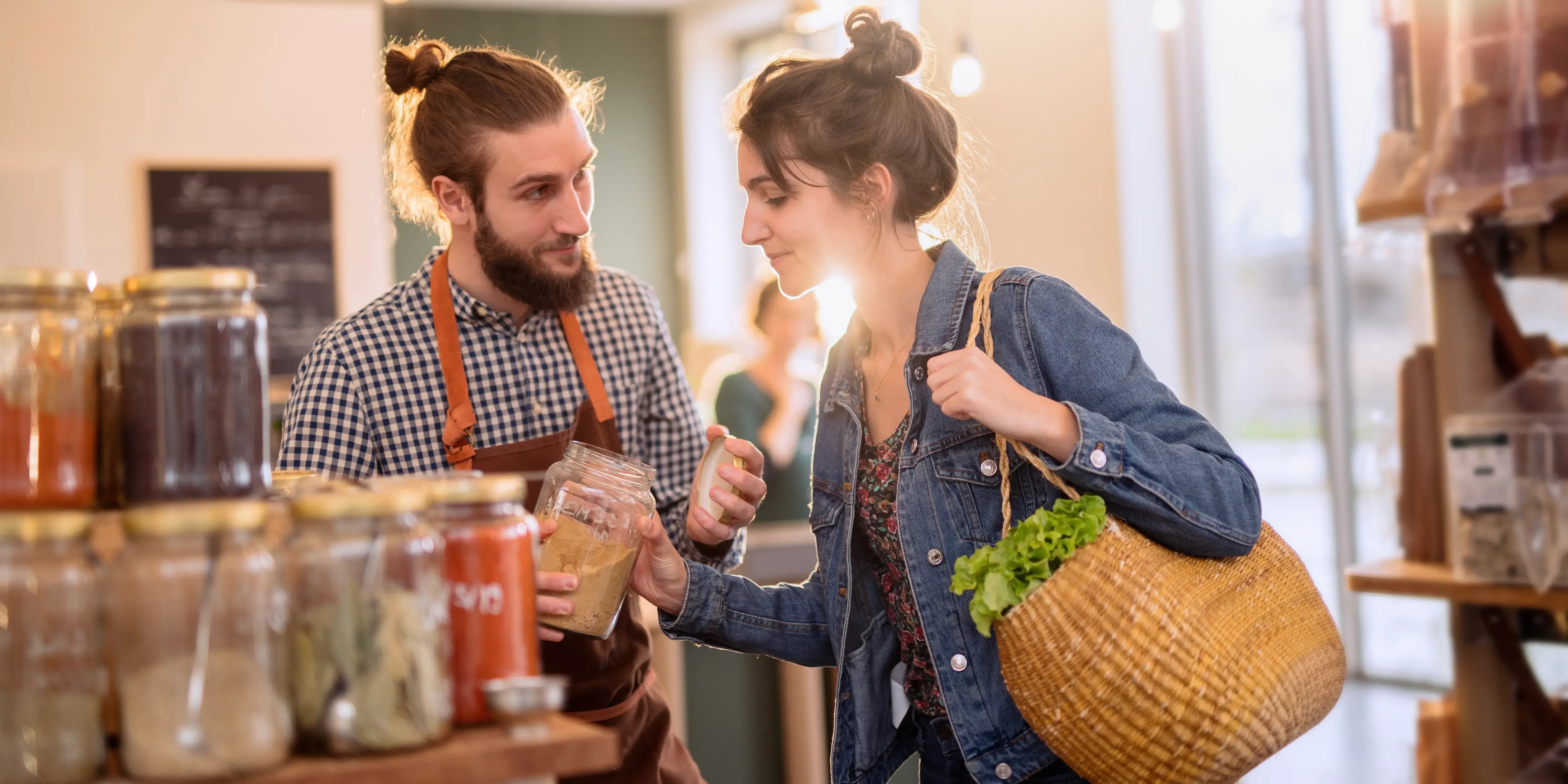 Mujer joven comprando en una tienda de comida a granel y el vendedor le aconseja comprar especias orgánicas en tarro como consumo consciente y vivir el lujo con estrategia