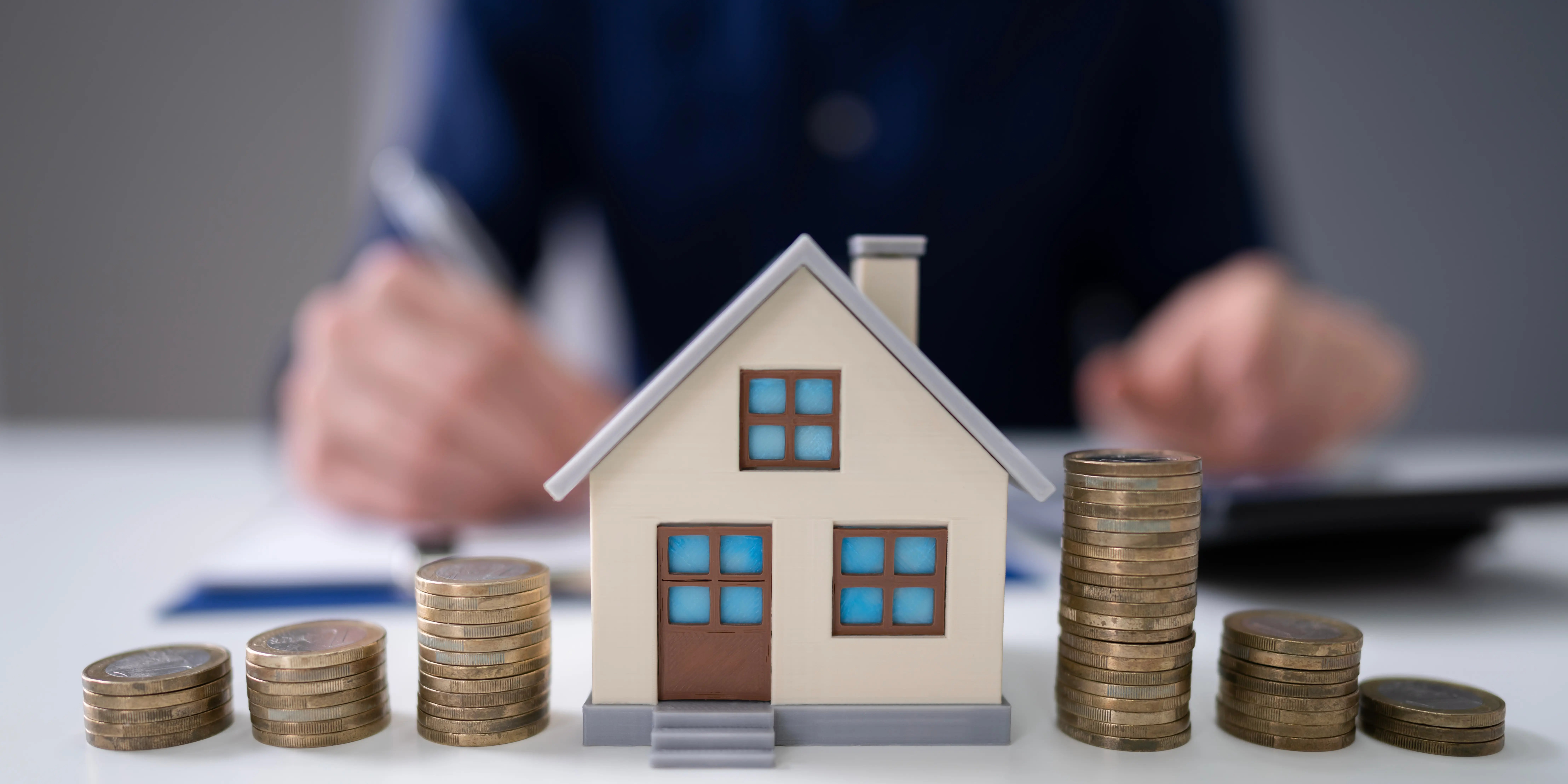 Torres de monedas al rededor de una casa de madera y al fondo un hombre calculando el impuesto a la plusvalía